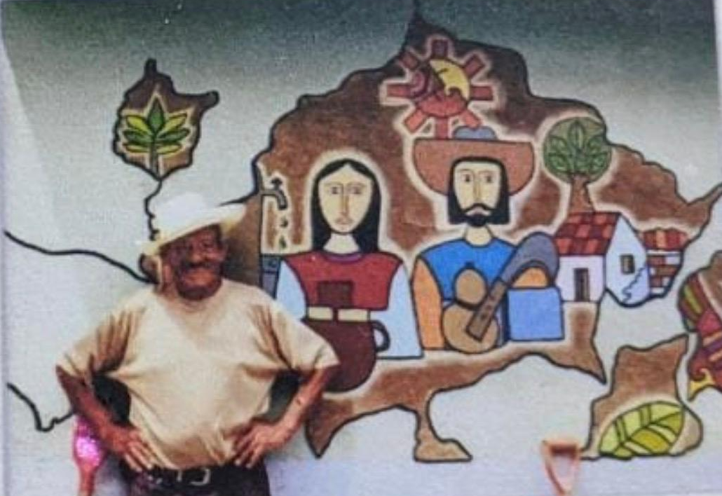 Papa Chico standing beside a mural reflecting the artistic roots of La Palma, El Salvador.